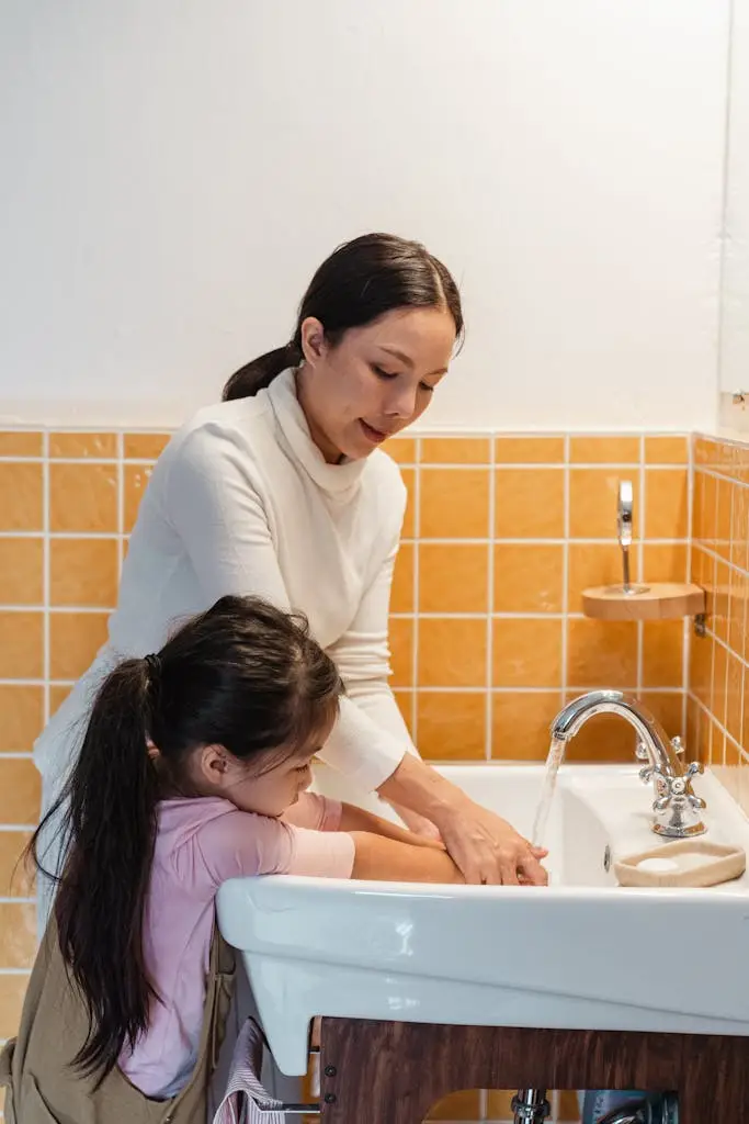 Side view of ethnic mother teaching adorable little daughter to wash hands in big white washing basin in modern bathroom